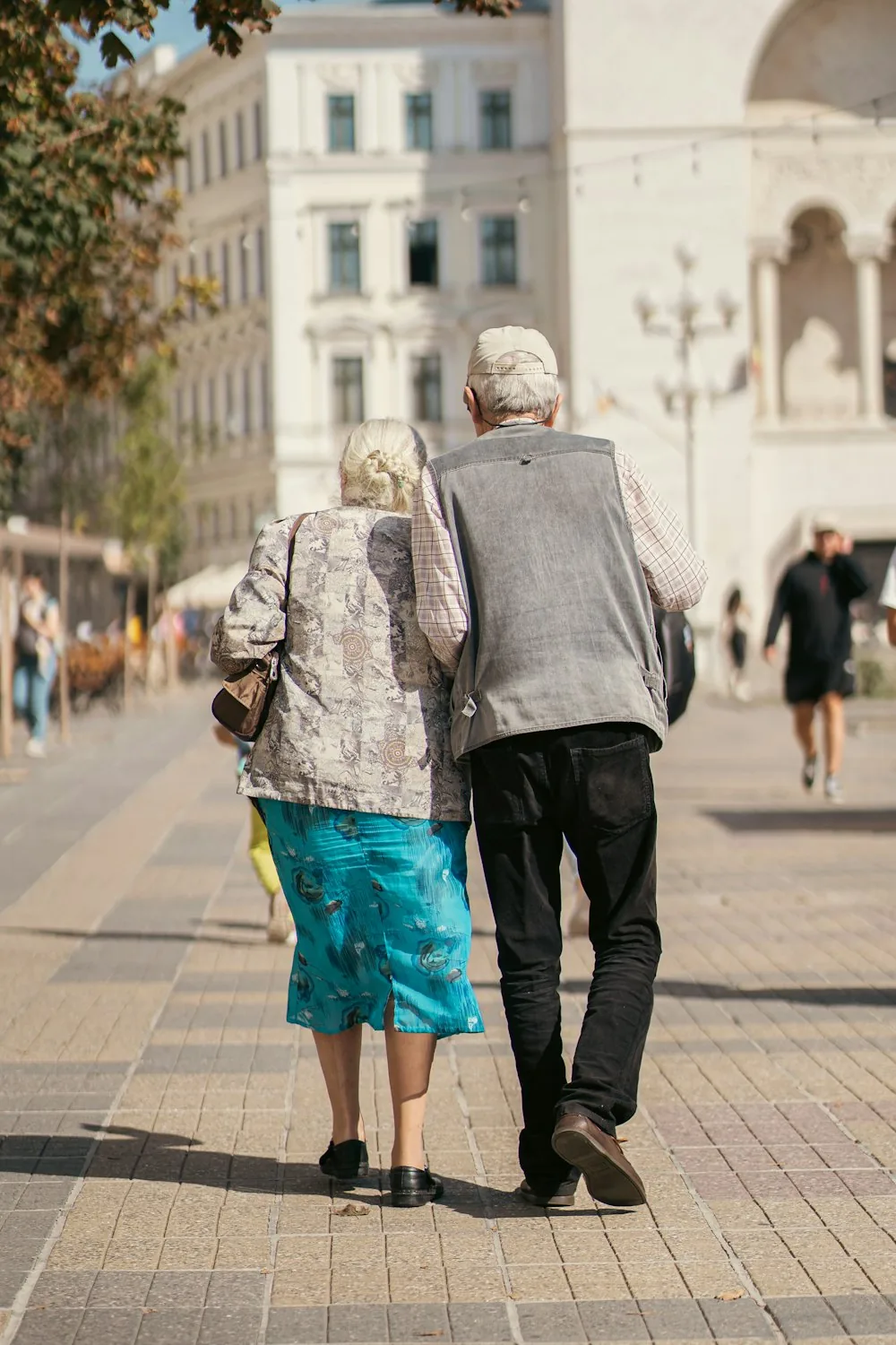 An older couple walking arm in arm along a sunlit pavement