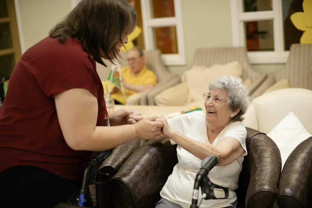A caregiver helping a smiling older woman up from her chair